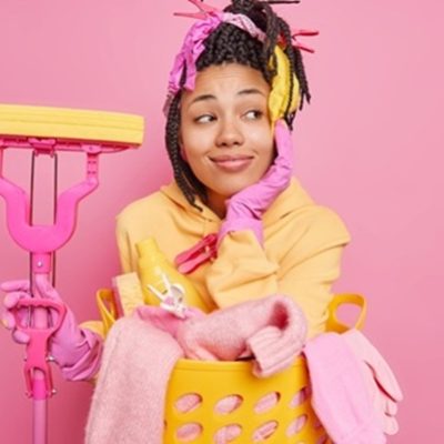 people housework and housekeeping concept. dreamy housewife keeps hand on face being deep in thoughts befores washing room holds mop poses near basket of laundry isolated on pink studio wall